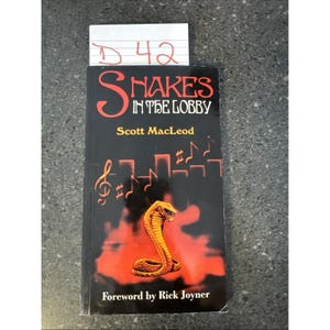 May include: A book titled "Snakes in the Lobby" by Scott MacLeod. The cover shows an orange cobra on a black background with musical notes and red accents. The foreword is by Rick Joyner.