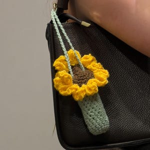 Puede incluir: Un charm de girasol tejido a crochet, con pétalos amarillos, centro marrón y tallo verde. El charm está sujeto a un bolso negro con un cordón verde claro. Un accesorio brillante y alegre.