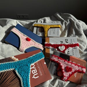 Könnte beinhalten: Eine Sammlung handgefertigter Häkel-String-Lesezeichen in verschiedenen Farben, darunter Rosa, Gelb, Rot und Türkis. Jedes Lesezeichen hat ein Herz oder eine dekorative Verzierung und liegt auf einem Buch.