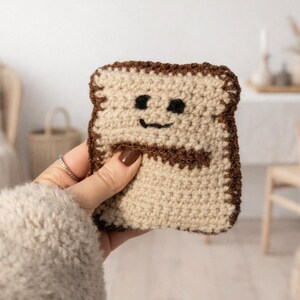 May include: Crocheted toast-shaped item with a smiling face. The toast is beige with brown details and a dark brown outline. The item is held by a person with a beige sweater and a silver ring.