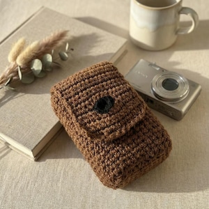 May include: A brown crocheted phone case with a flap and a black button closure. The case is handmade and sits on a neutral-colored surface next to a book, a mug, and a camera.
