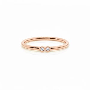 May include: Rose gold ring with two small, round, clear gemstones. The ring has a thin band and a polished finish. The gemstones are set close together, creating a delicate design. The ring is displayed on a white background.