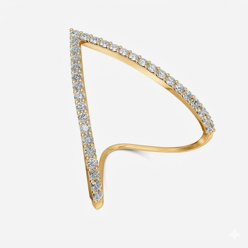 May include: A gold-colored ring with a unique, open design. The ring features a triangular shape lined with small, clear gemstones. The band curves around the finger, creating a modern and elegant look. The ring is set against a plain white background.