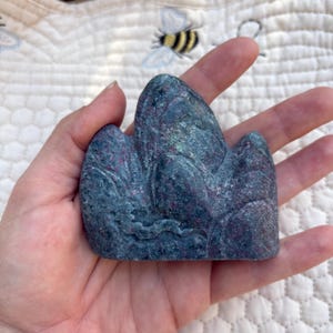May include: A hand holding a blue and purple stone carving of a mountain range. The stone has a textured surface with variations in color, including shades of blue, purple, and hints of green. The carving is detailed, with visible ridges and valleys.