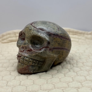May include: A carved stone skull with a detailed design. The skull is primarily gray with red and brown veining throughout. The skull is resting on a cream-colored, textured surface.