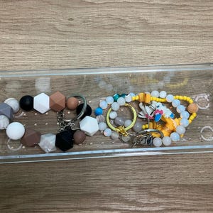 May include: A clear plastic container holding various beaded accessories. Items include bracelets with geometric and round beads in colors like white, black, and brown. There are also keychains and a yellow beaded necklace with a small figurine.