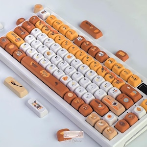 May include: A mechanical keyboard featuring a custom keycap set with a honey and bee theme. The keys are a mix of white, light orange, and dark orange, decorated with illustrations of bees, honey jars, flowers, and leaves. The spacebar displays the text Honey.