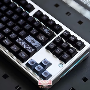 May include: A mechanical keyboard featuring a silver frame and black keycaps. The keycaps are decorated with white line art illustrations including snakes, celestial motifs, and an eye symbol. Standard function keys and navigation keys are present.