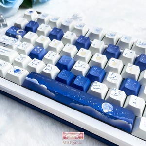 Op de afbeelding: Een wit en blauw mechanisch toetsenbord met aangepaste keycaps. De keycaps hebben een hemels thema met sterrenbeelden, planeten en een maanontwerp. De spatiebalk toont een nachtscène met de tekst "I was the moon".