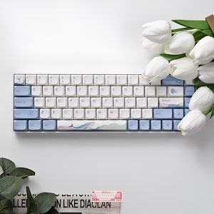 May include: A compact mechanical keyboard featuring a white and light blue color scheme. The spacebar displays a whale illustration, while other keys include icons of a sailboat, a jellyfish, and a cloud. The layout is standard QWERTY.