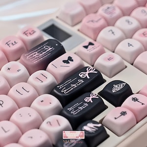 以下が含まれることがあります： ライトピンクとブラックのキーキャップを組み合わせたメカニカルキーボード。ピンクのキーには黒い記号、数字、リボンや鍵のアイコンが描かれています。黒いキーには白い筆記体の文字、リボンのイラスト、王冠のアイコンが施されています。