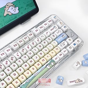May include: A mechanical keyboard with a transparent frame and gradient-colored keycaps in white, yellow, and green. The keys feature brown lettering and various cartoon animal illustrations, including a space bar with a landscape scene.