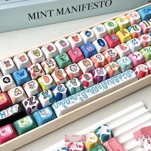 May include: A mechanical keyboard set featuring multi-colored keycaps with various cartoon illustrations, letters, and symbols. The spacebar has a blue dripping liquid design. The keys are arranged in a tray, with additional loose keycaps nearby.