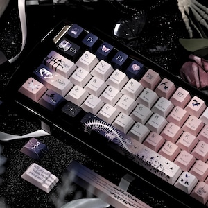 Puede incluir: Un teclado mecánico con un degradado de color que va del morado oscuro al rosa claro. Las teclas presentan tipografía gótica, ilustraciones de esqueletos y motivos de mariposas. La barra espaciadora muestra un gráfico detallado de una columna vertebral.
