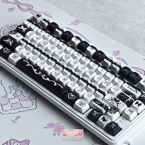 May include: A white and black mechanical keyboard with custom keycaps. The keycaps feature various designs, including bows, hearts, and lace patterns. The keyboard sits on a light pink and white patterned surface. The brand name "MAPStore" is visible.
