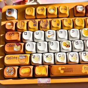 May include: A custom mechanical keyboard with honey-themed keycaps. The keycaps are a mix of white and yellow, featuring illustrations of honey pots, bees, and floral designs. The space bar reads "ORGANIC HONEY".