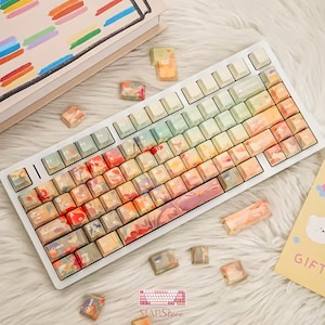 May include: A mechanical keyboard featuring a white frame and custom keycaps with a watercolor-style gradient pattern in shades of orange, red, yellow, and soft green. The keys display standard alphanumeric characters and function labels in white.