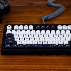 Puede incluir: Un teclado mecánico negro con teclas blancas y azul marino. El teclado presenta una distribución QWERTY estándar con teclas de función y una barra espaciadora decorada con un diseño espacial. El teclado está sobre una superficie de madera.