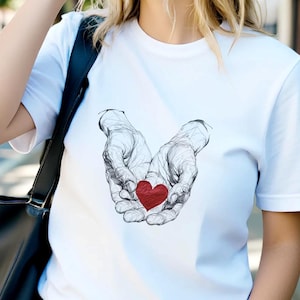 Può includere: Una t-shirt bianca a maniche corte con un disegno a linee nere di due mani a coppa che tengono un piccolo cuore rosso pieno. La maglietta è abbinata a jeans blu, una borsa a tracolla nera e occhiali da sole scuri sul viso.