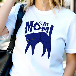 Könnte beinhalten: Weißes T-Shirt mit einer marineblauen Katzen-Grafik und dem Text "Cat Mom". Die Katzen-Grafik hat gelbe Augen. Die Person trägt eine Sonnenbrille und blaue Jeans. Eine schwarze Tragetasche wird über der Schulter getragen.