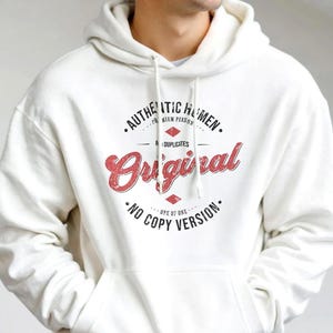 Könnte beinhalten: Cremefarbener Kapuzenpullover mit Fronttasche und Kordelzug. Der Kapuzenpullover zeigt den Text "AUTHENTIC HUMAN, ORIGINAL, NO COPY VERSION" in einem rot-schwarzen Design. Die Person trägt eine graue Jogginghose.