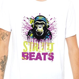 Op de afbeelding: Wit T-shirt met een kleurrijke afbeelding van een aap met een koptelefoon, met verfspatten en de tekst "STREET BEATS" in een gestileerd lettertype. Het ontwerp bevat paarse, groene en gele accenten.