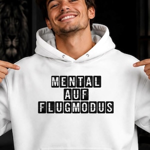 Op de afbeelding: Witte hoodie met een voorzak en een trekkoord. De tekst "MENTAL AUF FLUGMODUS" staat in zwarte blokletters op de borst. De persoon op de foto wijst naar de tekst.