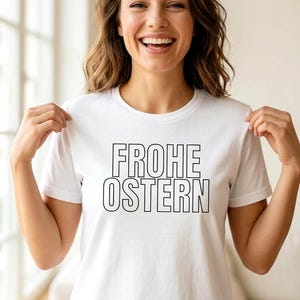 Könnte beinhalten: Weißes T-Shirt mit dem deutschen Text "FROHE OSTERN" in schwarzen Blockbuchstaben. Das T-Shirt wird von einer Person mit braunen Haaren und einem Lächeln hochgehalten. Das Shirt ist ein lässiger Stil.