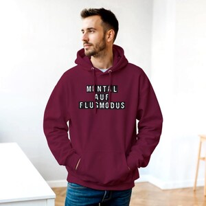 Op de afbeelding: Een bordeauxrode hoodie met een wit trekkoord en een voorzak. De hoodie heeft de Duitse tekst "MINTAL AUF FLUGMODUS" in witte letters op een zwarte achtergrond. De persoon draagt een blauwe spijkerbroek.