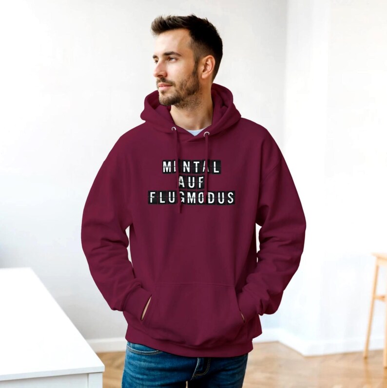 Op de afbeelding: Een bordeauxrode hoodie met een wit trekkoord en een voorzak. De hoodie heeft de Duitse tekst "MINTAL AUF FLUGMODUS" in witte letters op een zwarte achtergrond. De persoon draagt een blauwe spijkerbroek.