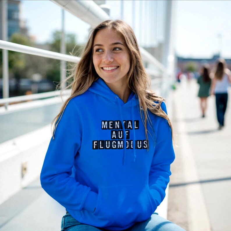 Op de afbeelding: Een felblauwe hoodie met de tekst "MENTAL AUF FLUGMODUS" in een zwart-wit blokletterontwerp. De hoodie heeft een voorzak en een capuchon. De persoon draagt de hoodie en glimlacht.