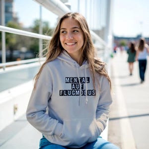 Op de afbeelding: Een lichtgrijze hoodie met de tekst "MENTAL AUF FLUGMODUS" in zwarte blokletters. De hoodie heeft een voorzak en een capuchon. De persoon draagt een blauwe spijkerbroek.
