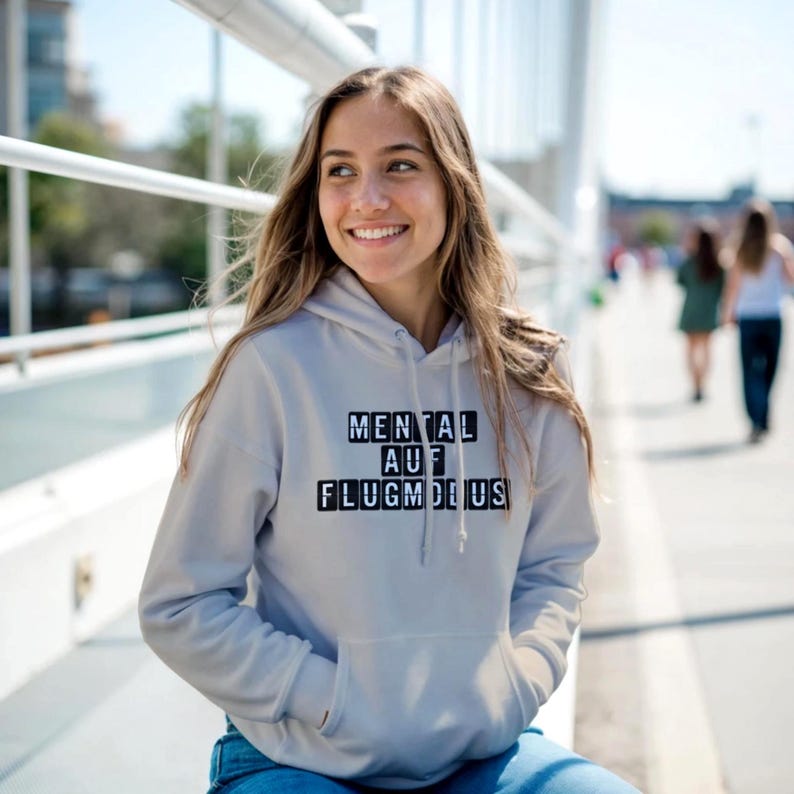 Op de afbeelding: Een lichtgrijze hoodie met de tekst "MENTAL AUF FLUGMODUS" in zwarte blokletters. De hoodie heeft een voorzak en een capuchon. De persoon draagt een blauwe spijkerbroek.