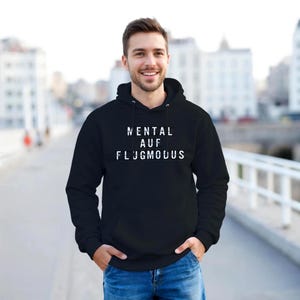 Op de afbeelding: Een zwarte hoodie met de witte tekst "MENTAL AUF FLUGMODUS". De sweatshirt heeft een capuchon met trekkoord en een voorzak. De persoon op de foto lacht en draagt een blauwe spijkerbroek.