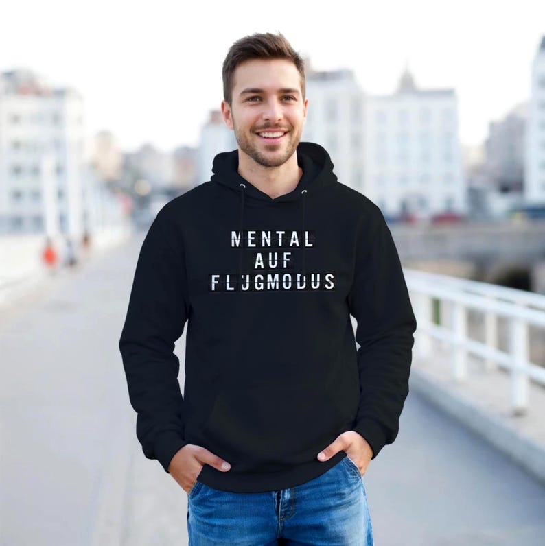 Op de afbeelding: Een zwarte hoodie met de witte tekst "MENTAL AUF FLUGMODUS". De sweatshirt heeft een capuchon met trekkoord en een voorzak. De persoon op de foto lacht en draagt een blauwe spijkerbroek.