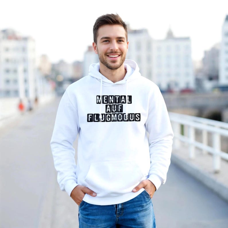 Op de afbeelding: Witte hoodie met de tekst "MENTAL AUF FLUGMODUS" in zwarte blokletters. De hoodie heeft een capuchon met trekkoord en een voorzak. De persoon draagt een blauwe spijkerbroek.