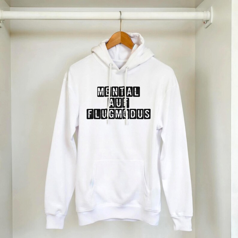 Op de afbeelding: Witte hoodie met een voorzak en een capuchon met trekkoord. De hoodie toont de tekst "MENTAL AUF FLUGMODUS" in zwarte blokletters. De hoodie hangt aan een houten hanger.