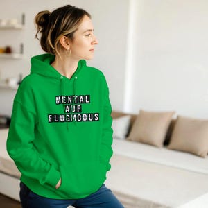 Op de afbeelding: Een felgroene hoodie met de Duitse tekst "MENTAL AUF FLUGMODUS" in zwarte blokletters. De hoodie heeft een capuchon en een voorzak. De persoon draagt de hoodie.