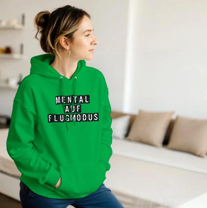 Op de afbeelding: Een felgroene hoodie met de Duitse tekst "MENTAL AUF FLUGMODUS" in zwarte blokletters. De hoodie heeft een capuchon en een voorzak. De persoon draagt de hoodie.