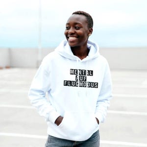 Op de afbeelding: Een witte hoodie met een voorzak en een trekkoord. De tekst "MENTAL AUF FLUGMODUS" is gedrukt in zwarte blokletters. De persoon op de foto lacht en draagt de hoodie.