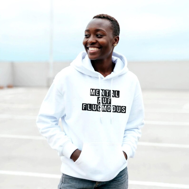 Op de afbeelding: Een witte hoodie met een voorzak en een trekkoord. De tekst "MENTAL AUF FLUGMODUS" is gedrukt in zwarte blokletters. De persoon op de foto lacht en draagt de hoodie.