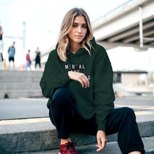 Op de afbeelding: Een donkergroene hoodie met de tekst "MENTAL" boven "THE" en "S" in het wit. De hoodie wordt gedragen met een zwarte broek en rode schoenen. De persoon zit op betonnen trappen.