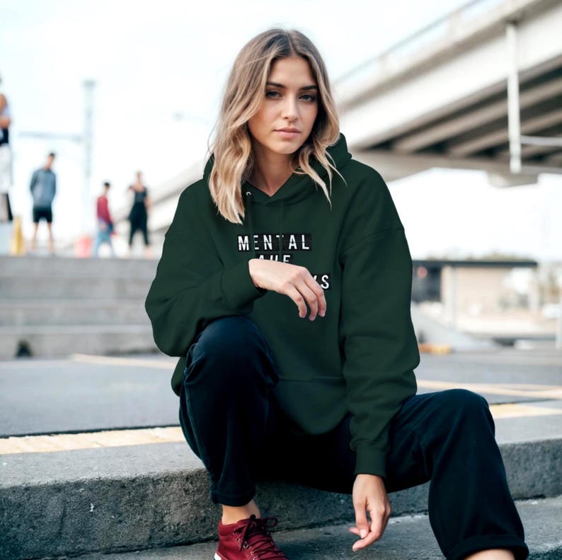 Op de afbeelding: Een donkergroene hoodie met de tekst "MENTAL" boven "THE" en "S" in het wit. De hoodie wordt gedragen met een zwarte broek en rode schoenen. De persoon zit op betonnen trappen.
