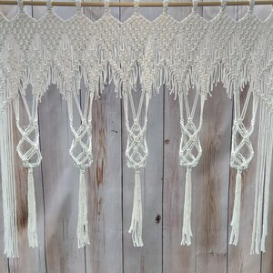 May include: A white macrame curtain with a geometric design. The curtain is suspended from a wooden rod and features intricate knotting patterns, with hanging tassels and decorative plant holders. The curtain is made of a soft, natural fiber.