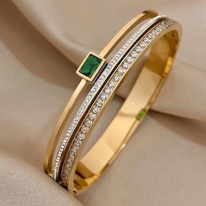 Può includere: Un bracciale rigido dorato con tre fasce. Una fascia è adornata con cristalli trasparenti, e un'altra presenta una gemma verde smeraldo quadrata. Il bracciale è posto su uno sfondo neutro.