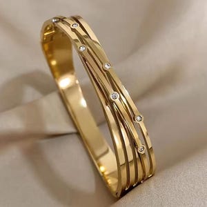 May include: A gold-coloured bangle bracelet with a layered design. The bracelet has multiple parallel bands, some of which are decorated with small, round, clear gemstones. The bracelet is shown on a neutral-coloured, slightly textured surface.