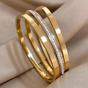 May include: Three gold-colored bangles. One bangle is adorned with a row of small, clear crystals. The bangles are stacked together, creating a layered look. The jewelry has a shiny, reflective surface.