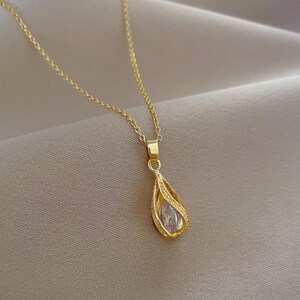 Op de afbeelding: Een goudkleurige ketting met een fijne schakelketting en een druppelvormige hanger. De hanger bestaat uit een goudkleurig kooiframe met textuurdetails, waarin een enkele, heldere, marquise-geslepen edelsteen is geplaatst.