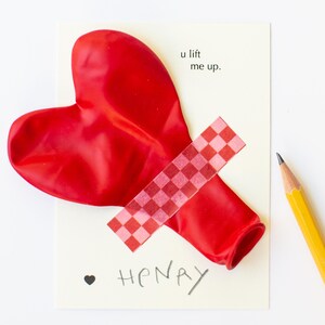 Puede incluir: Un globo rojo en forma de corazón está pegado a una tarjeta con cinta adhesiva rosa a cuadros. La tarjeta tiene el nombre escrito a mano "Henry" y la frase "u lift me up". Un lápiz amarillo está en el marco.