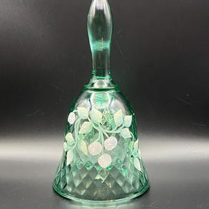 May include: A green glass bell with a long handle and a diamond-patterned base. The bell is decorated with white and pink floral designs. The bell is a decorative item.